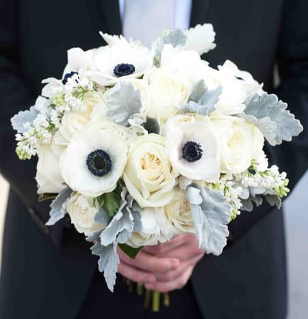 In questa foto, uno sposo tiene in mano un bouquet invernale sui toni del bianco realizzato con anemoni, rose e bouvardia 