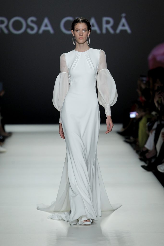 In questa foto la modella indossa un abito da sposa semplice 2023 con maniche trasparenti  Rosa Clarà