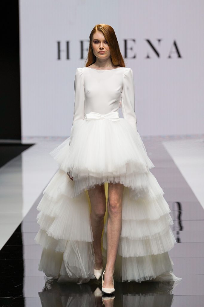 In questa foto la modella indossa un abito da sposa corto con maniche lunghe e gonna di tulle con coda a balze di Maison Signore 2023.