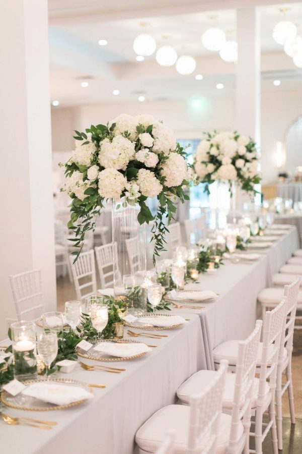 In questa foto addobbi per tavoli in stile elegante con alzatine di rose e ortensie bianche disposte su un tavolo imperiale con tovagliato colore perla e mise en place colore oro e cristallo