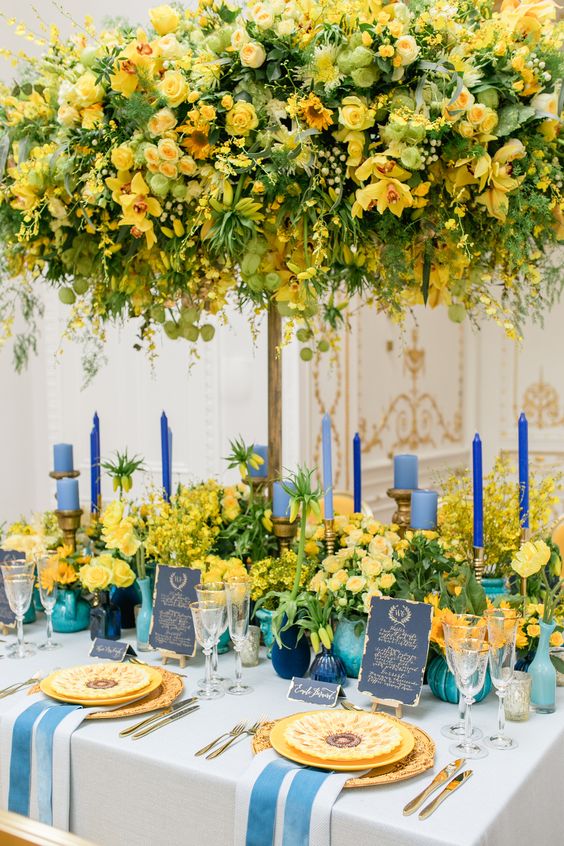 In questa foto una mise en place nei toni dell'azzurro, del blu e del giallo. l centro è presente un grande centrotavola a forma di albero con. fiori gialli. Sul tavolo sono presenti altre composizioni abbinate, candele di diverse dimensioni di colore azzurro e blu e piatti gialli decorati con nastri a contrasto