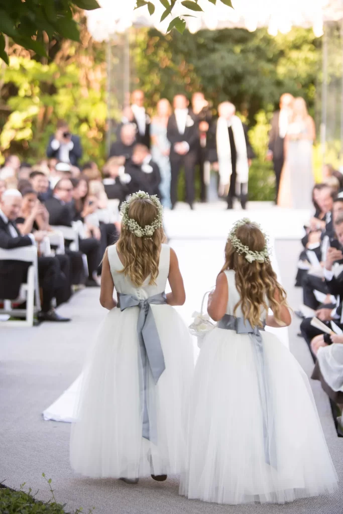 In questa foto due damigelle fotografate di spalle fanno il loro ingresso alla cerimonia. Indossano un abito lungo con gonna in tulle, un fiocco colore azzurro carta da zucchero e una coroncina di gypsophila bianca