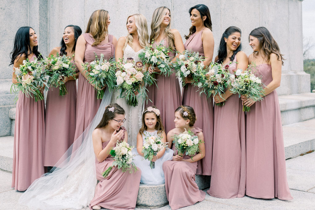 In questa foto una sposa posa con le sue damigelle adulte e le piccole damigelle. Tutte indossano un abito lungo di colore rosa ciclamino e tengono tra le mani un bouquet uguale a quello della sposa