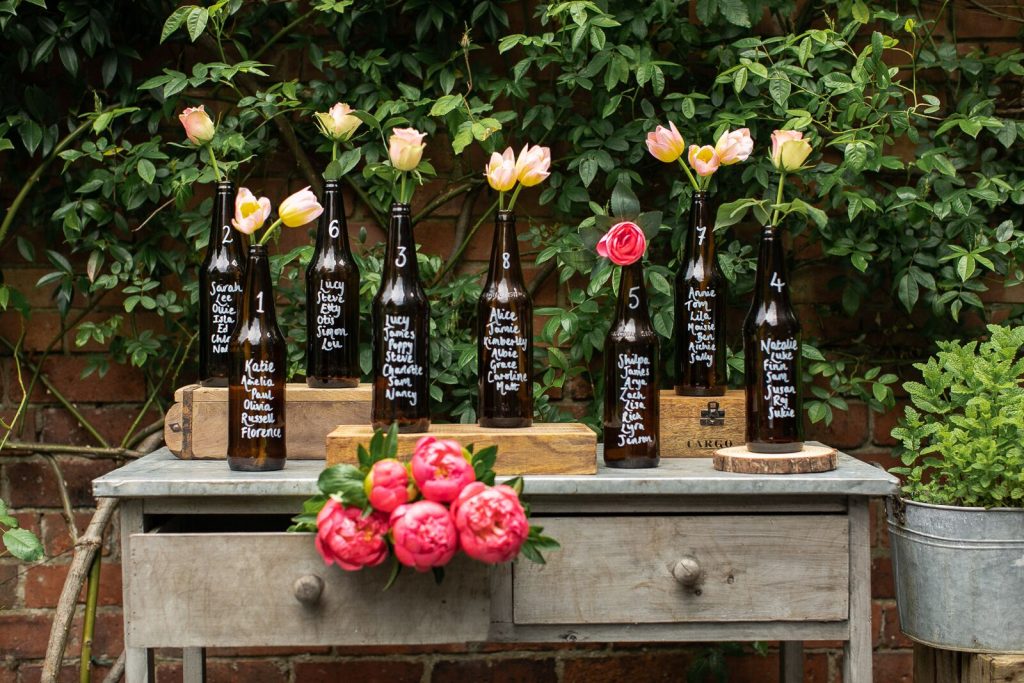 In questa foto un tableau di matrimonio fai da te creato con bottiglie di vetro riciclate dipinte con i nomi degli invitati e decorate con rose e tulipani. tutte le bottiglie sono sistemate su un'antica scrivani