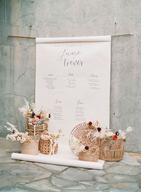 In questa foto un tableau di matrimonio composto da un grande poster di colore bianco con i nomi degli sposi e gli elenchi dei tavoli. Alla base sono presenti 6 ceste di paglia piene di fiori nei colori del rosso, rosa, bianco e bordeaux