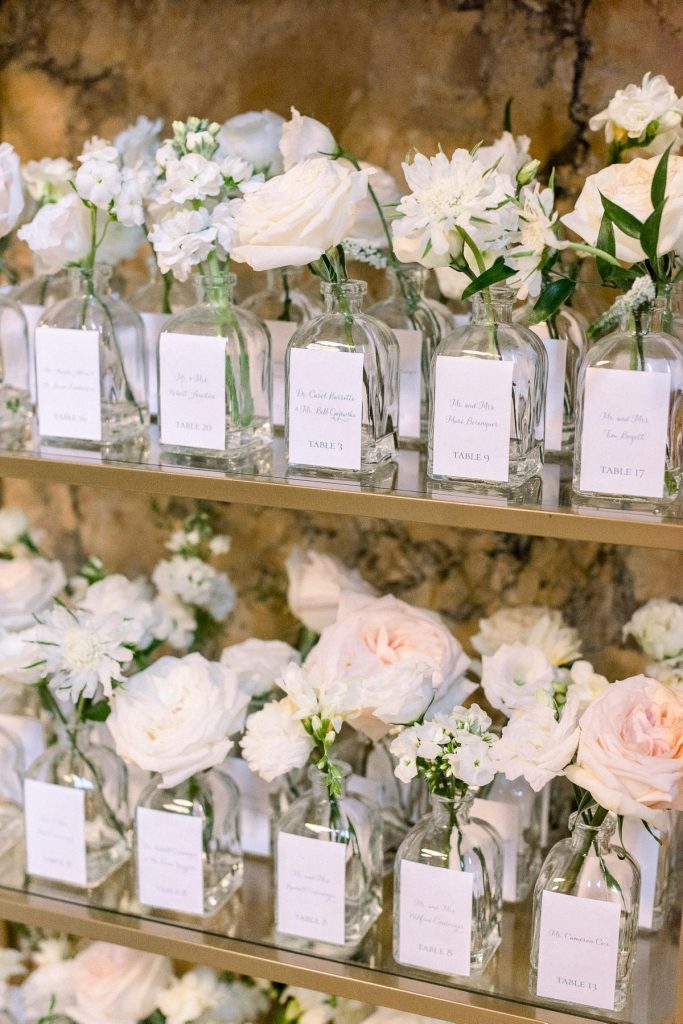 In questa foto una tableau matrimonio romantico con vasetti di vetro in cui sono inseriti boccioli di rose di varietà diverse tutti distribuiti su mensole di vetro