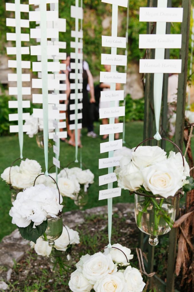 In questa foto un tableau di nozze appeso all'albero con piccoli bigliettini e basi di rose di colore bianco