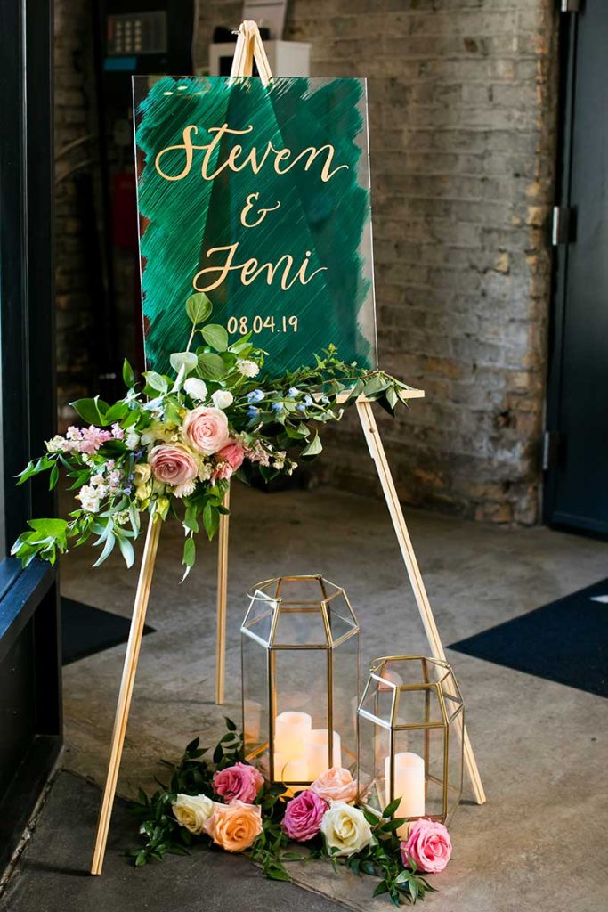 In questa foto un tableau welcoma mariage di plexiglass pennellato di verde scuro poggiato su un treppiedi decorata con rose, fiori e foglie