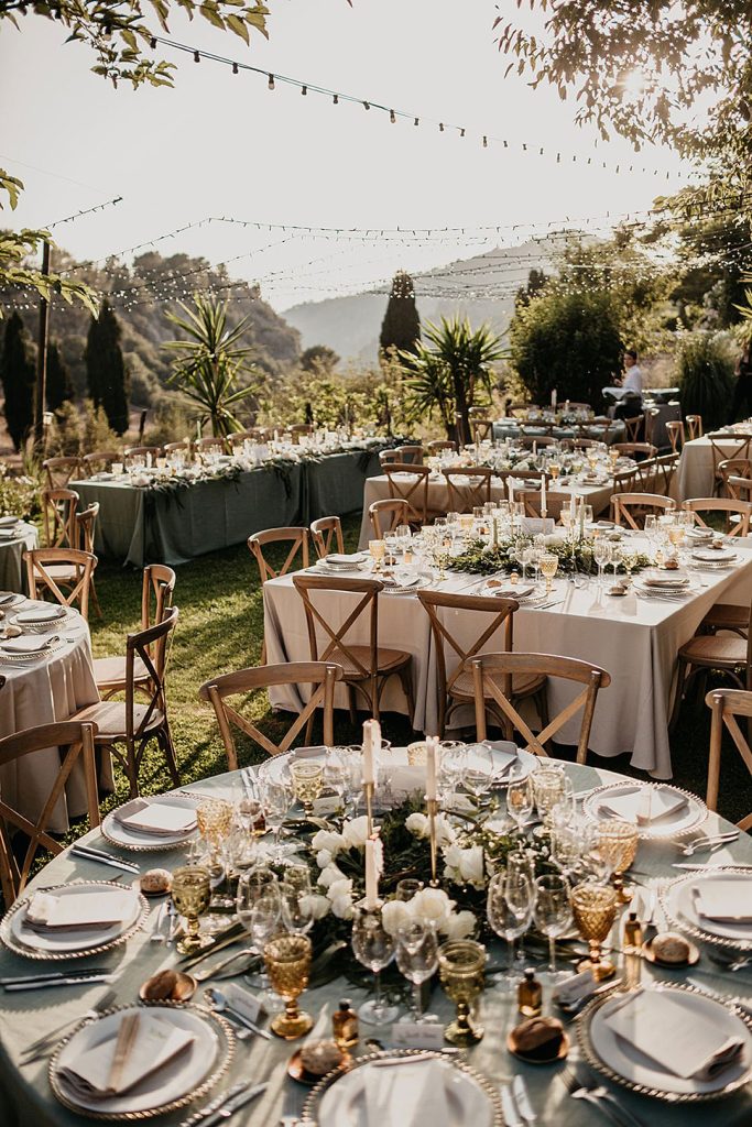 In questa foto tavoli rotondi e quadrati allestiti in stili Rustic Chic con tovagliato bianco e verde salvia e mise en place colore oro