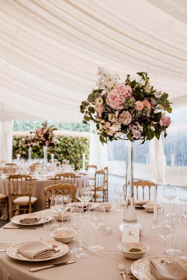 In questa foto un ricevimento con tavoli per matrimonio rotondi allestiti in stile elegante con tovagliato color tortora e centrotavola in cristallo con fiori rosa