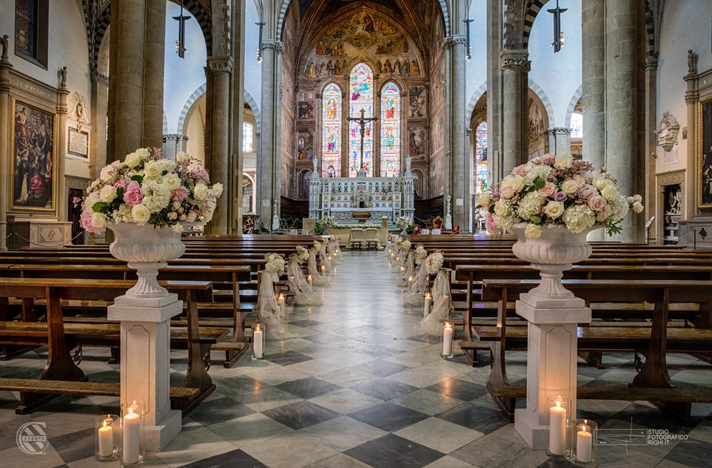 In questa foto un meraviglioso allestimento a cura di Corsini Events. Fiori bianchi, in varie sfumature di rosa e lilla su di eleganti e candide strutture per un ingresso in chiesa raffinato ed indimenticabile.  Vicino alle panche ci sono anche candele e morbidi tessuti bianchi. 