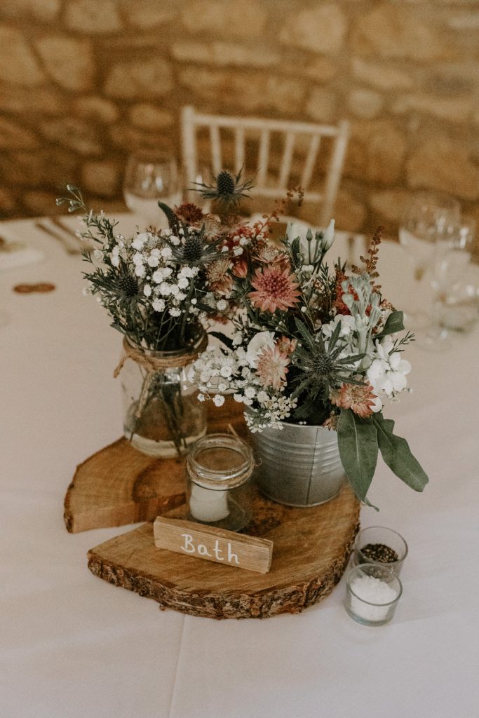 In questa foto un'idea di centrotavola country chic con fiori di campo sistemati in un vaso trasparente e un secchiello di metallo poggiati su una base in legno