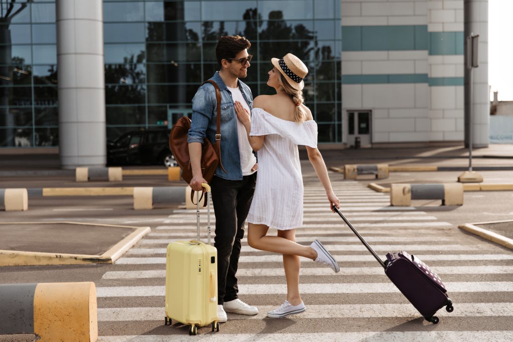 In questa una coppia sulle strisce pedonali prima di entrare in aeroporto per il loro viaggio di nozze. Lei tiene una mano poggiata sul petto di lui e una gamba alzata. Entrambi tengono con una mano un trolley