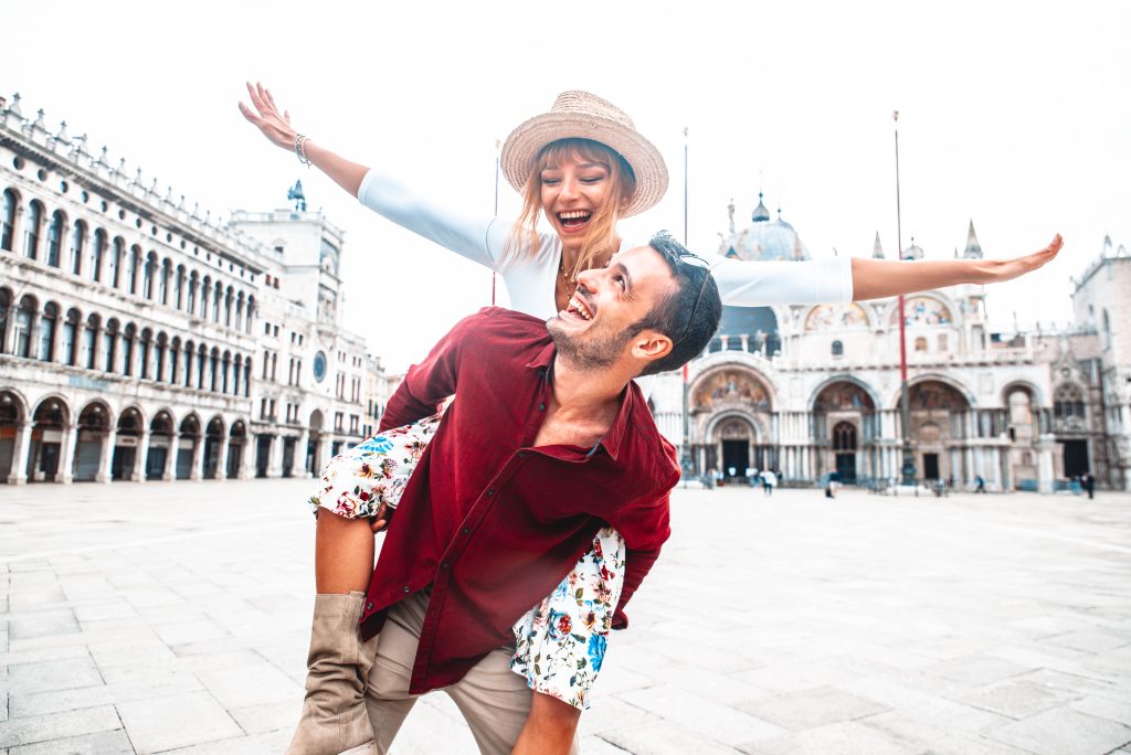 In questa foto una coppia a Piazza San Marco a Venezia. Lei sulle spalle di lui sorride fingendo di planare con le braccia. Lui la guarda e sorride