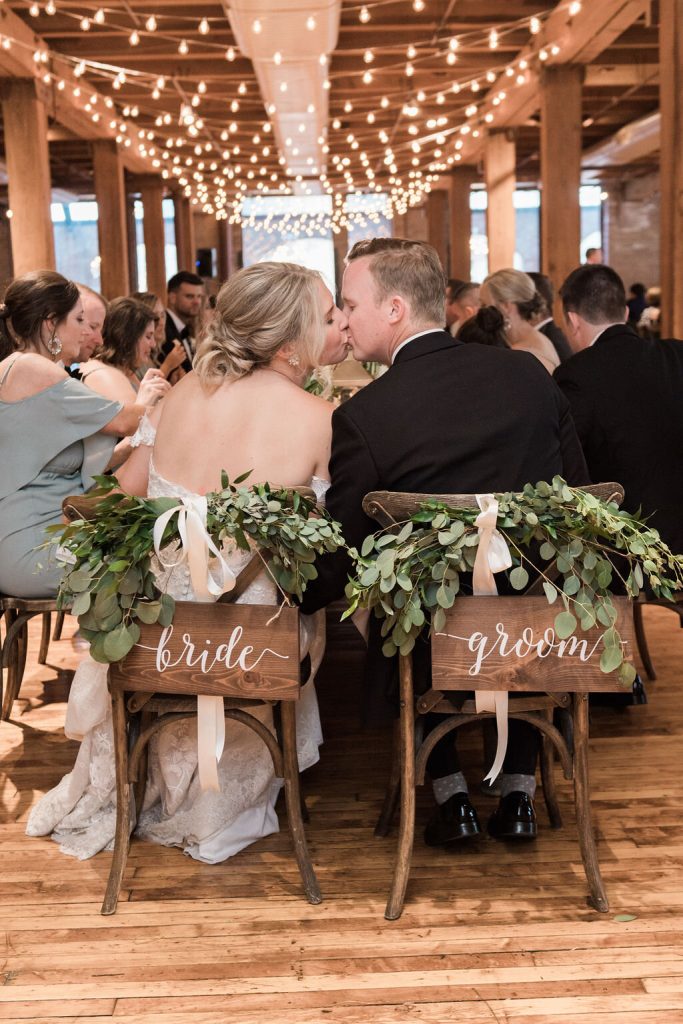 In questa foto due sposi di spalle seduti su sedie decorate con foglie e due banner di legno con scritto Bride e Groom