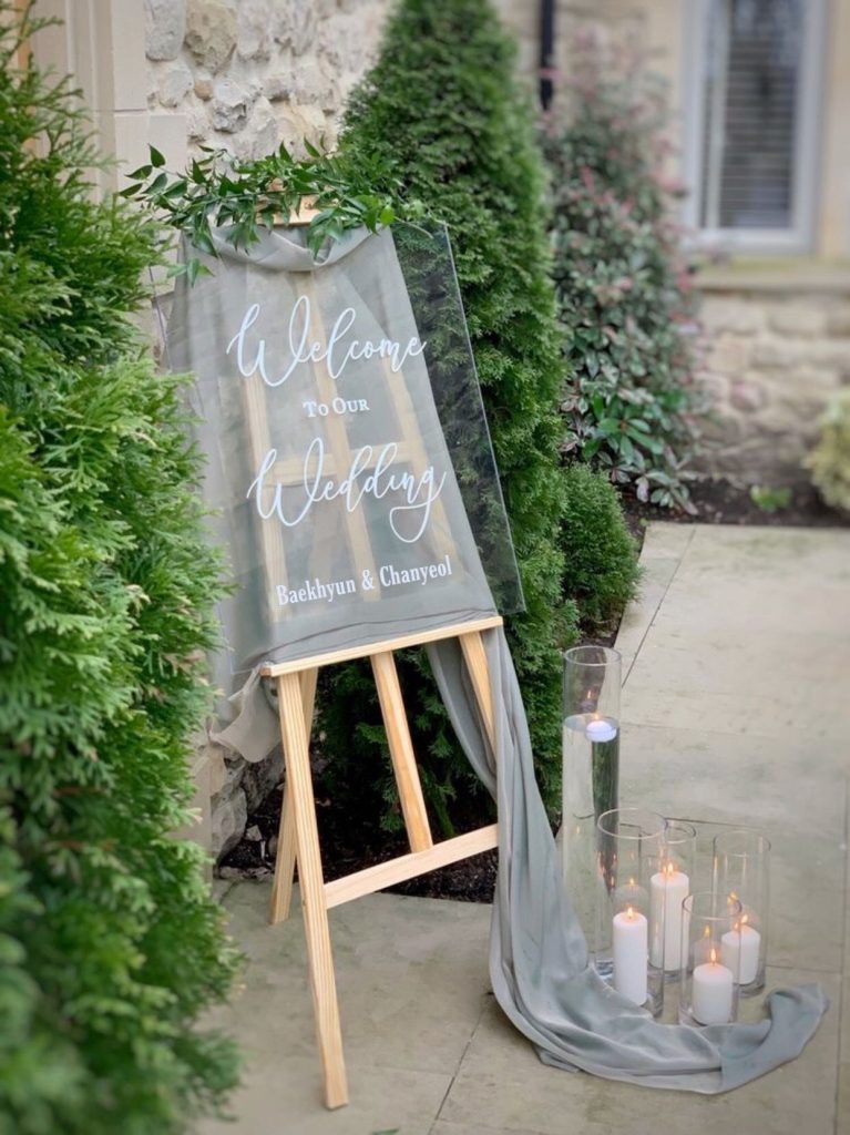 In questa foto un'idea di welcome board in plexiglass abbinato a un drappo di stoffa colore grigio perla e candele colore bianco disposte dentro portacandela trasparenti