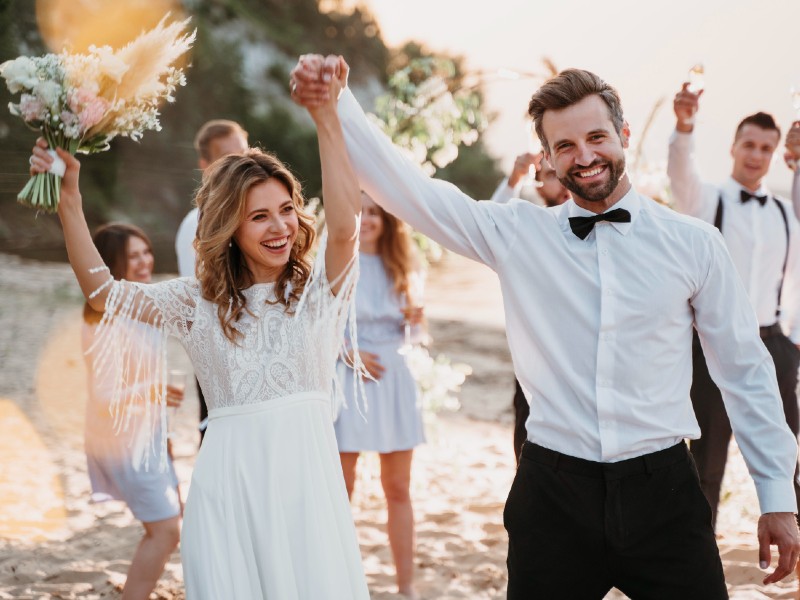 In questa foto di idee per matrimonio, due sposi camminano sulla spiaggia con i loro amici. Gli sposi si tengono per mano che alzano, la sposa tiene il suo bouquet con la mano destra alzata