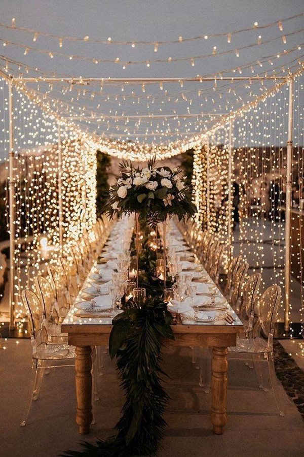 In questa foto un tetto di luci su un tavolo imperiale di legno come idee per decorazioni matrimonio. Al centro del tavolo è presente un runner di foglie mentre ai lati sono presenti sedie trasparenti