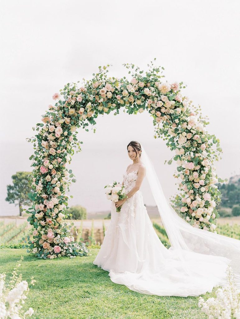 In questa foto una sposa con abito ad A decorata con ramage floreali posa su un prato con il suo bouquet davanti ad un arco di rose colore bianco e rosa