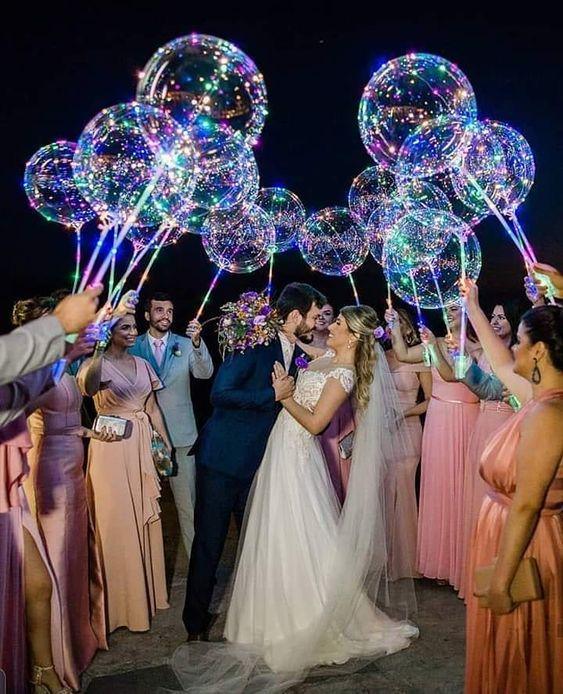 In questa foto due sposi circondati dai loro invitati che tengono in mano palloncini trasparenti con luci al led