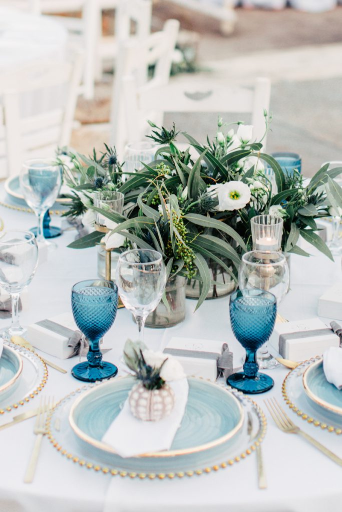 In questa foto un'idea di allestimento a tema mare nei toni del blu e del bianco. I piatti e i bicchieri sono di colore blu e i fiori di colore bianco. Ricci e conchiglie sono usati come segnaposto