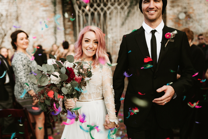 In questa foto idee matrimonio per l'uscita dalla chiesa: due sposi sorridenti camminano tra piume colorate lanciate dagli invitati
