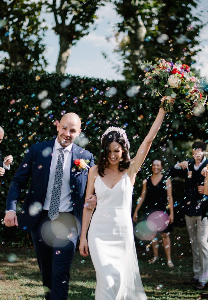 In questa foto idee matrimonio per l'uscita dalla chiesa: due sposi camminano tra una nuvola di bolle di sapone. la sposa alza vittoriosa il bouquet con la mano sinistra
