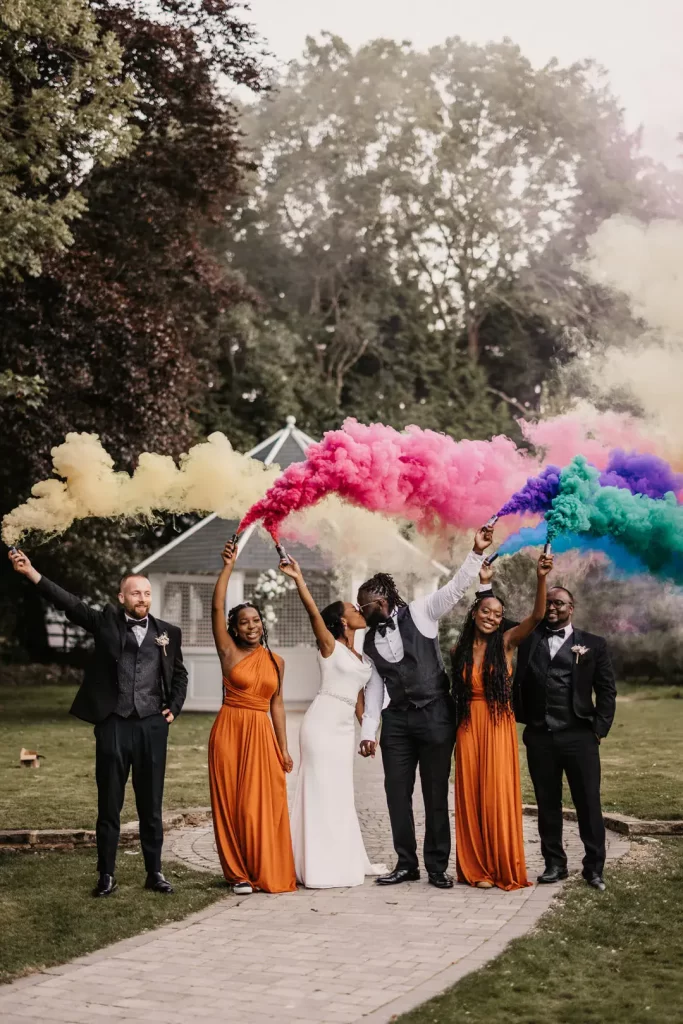 In questa foto idee matrimonio per l'uscita dalla chiesa: due sposi che si baciano tengono in mano fumogeni colorati circondati da damigelle e testimoni