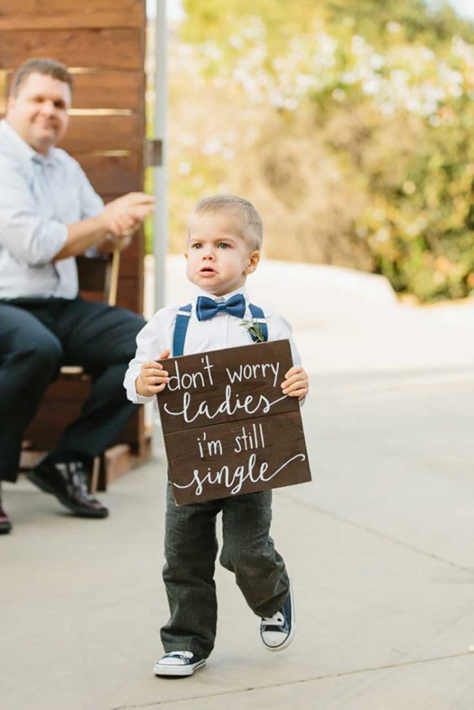 In questa foto idee matrimonio fai da te: un paggetto porta un cartello di legno con scritto "Don't worry ladies, i'm still single"