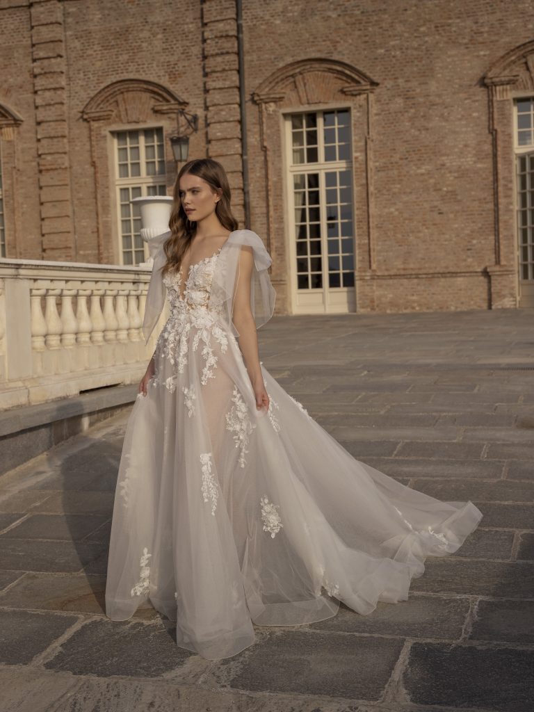 In questa foto un modello di abiti da sposa Capri 2023 con fiocchi sulle spalle e decorazioni in pizzo