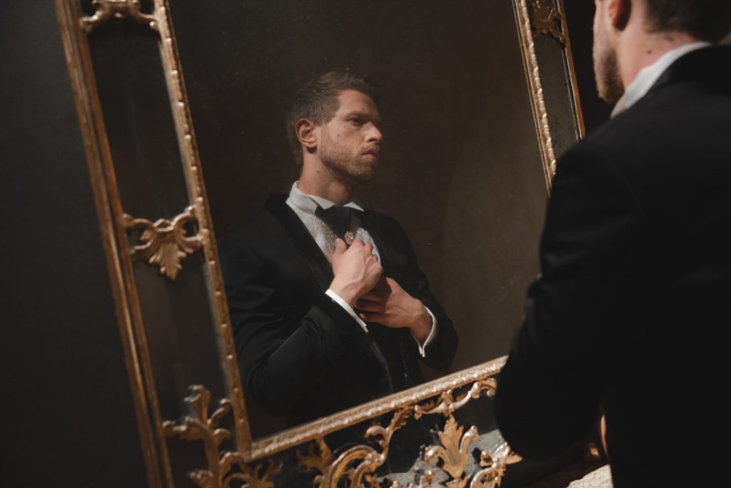 In questa foto un modello davanti a uno specchio si guarda mentre indossa un abito da sposo nero della collezione Kigili 2023