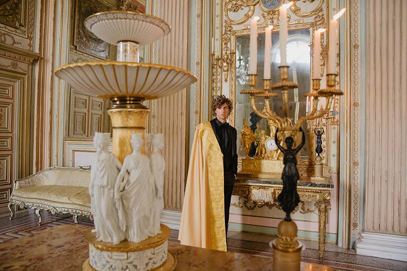 in questa foto un modello indossa un abito da sposo nero e sulle spalle un mantello in tessuto damascato color oro