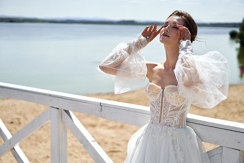 In questa foto una modella con la testa leggermente sollevata verso l'alto indossa uno degli abiti da sposa Annais Bridal 2023 in tulle colore bianco, corpetto a cuore con dettagli sparkling e maniche balloon rimovibili