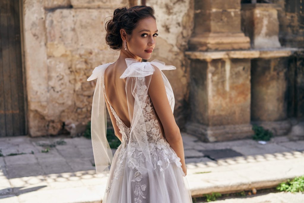 In questa foto una modella posa di spalle ruotando la testa verso destra e mostra un abito da sposa Annais Bridal 2023 con schiena scoperta, busto in pizzo floreale e fiocchi di tulle sulle spalle
