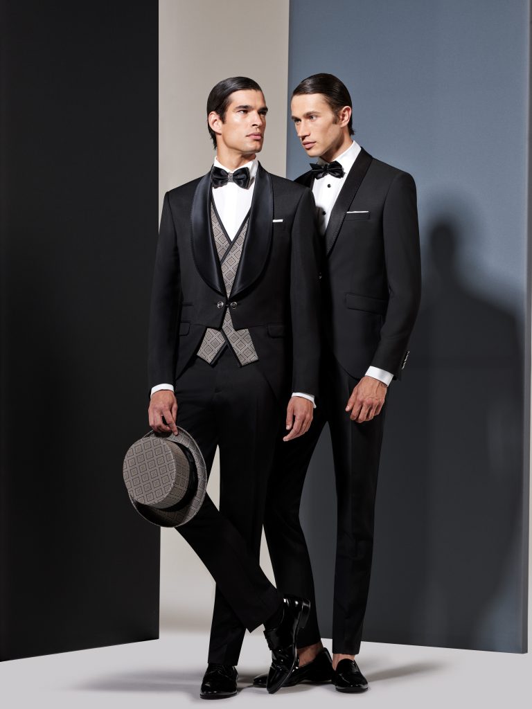 In questa foto due modelli indossano un frac e uno smoking di colore nero di Andrea Versali. Il modello in primo piano tiene nella mano destra un cilindro grigio abbinato al gilet