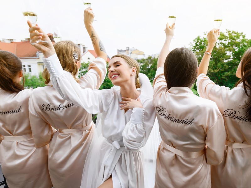 In questa foto una sposa festeggia il suo addio al nubilato con le sue amiche. Sono tutte affacciate su un balcone e tengono in alto un flute di champagne; le amiche sono girate di spalle e mostrano la scritta Bridesmaid sulla vestaglia. La sposa indossa una vestaglia bianca e un velo