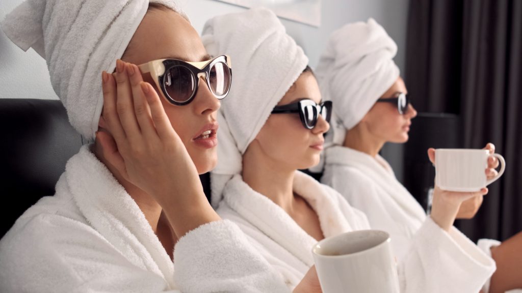 In questa foto tre ragazze in una spa con in accappatoio, occhiali da sole e capelli raccolti in un turbante. Tengono in mano un tazza bianca