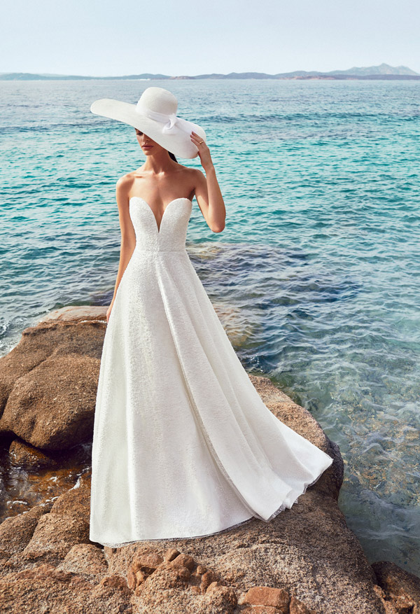 In questa foto una sposa su uno scoglio con il mare di sfondo. Indossa un abito broccato con profondo scollo a cuore un cappello bianco a falda larga con fiocco