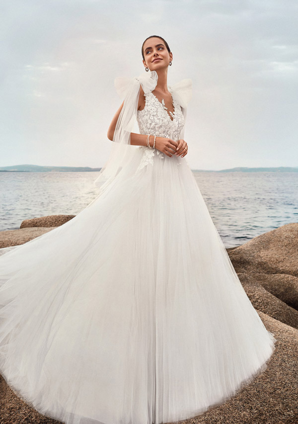 In questa foto una sposa su uno scoglio guarda verso l'alto e indossa un abito ad A in tulle con corpetto ricamato di perline e fiocchi sulle spalle