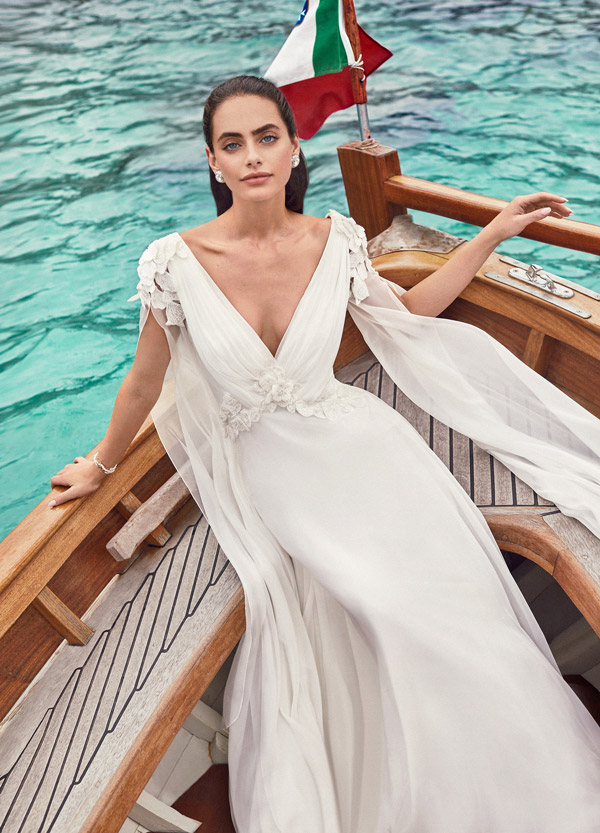 In questa foto una sposa su una barca  indossa un abito a sirena con scollo a V drappeggiato, dettagli floreali sulle spalle e sulla vita e maniche ampie ad ali