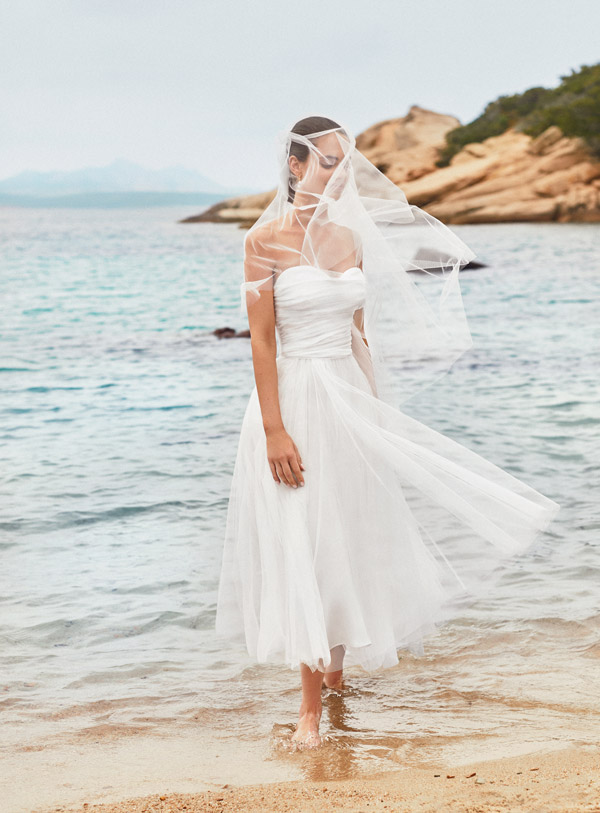 In questa foto una sposa sulla spiaggia con i piedi in acqua indossa un abito corto alle caviglie in tulle con corpetto drappeggiato con scollo a cuore