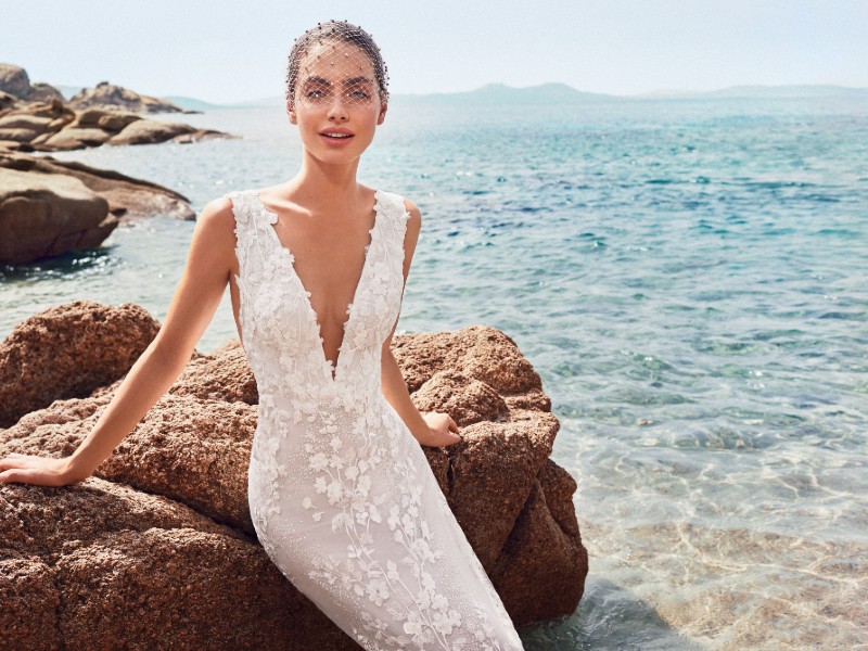 In questa foto una modella indossa un abito da sposa a sirena della collezione Atelier Emé 2023. La modella è seduta su un scoglio in riva al mare sullo sfondo