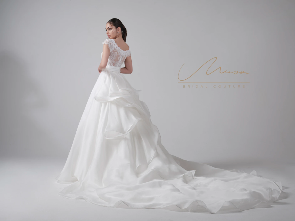 In questa foto una modella di spalle indossa un abito ampio con lunga coda di ruches in organza di Musa Bridal