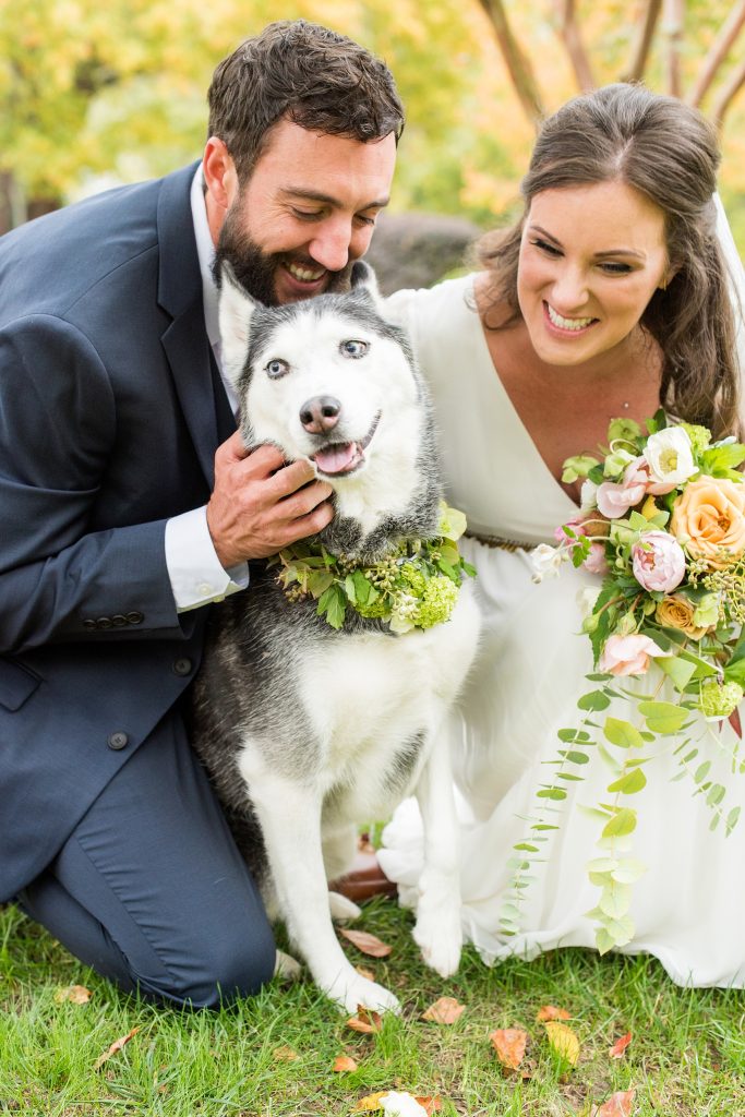 In questa foto due sposi leggermente chinati posano e abbracciano il loro cane sorridenti con l'aiuto di un Wedding Dog Sitter