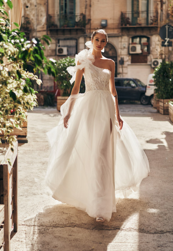 In questa foto una modella cammina indossando un abito monospalla della collezione 2023 del brand Annais Bridal