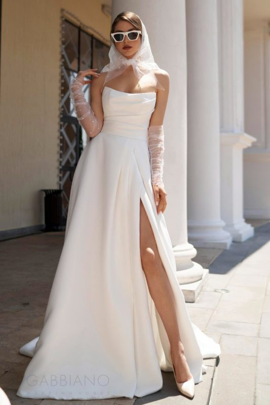 In questa foto una modella indossa un abito da sposa con spacco della collezione Gabbiano 2023. A completare il look, gli occhiali da sole
