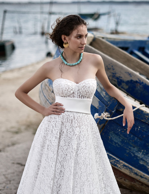 In questa foto una modella con il braccio poggiato su una barca mentre indossa un abito da sposa con corpetto bustier a cuore in pizzo all over, della collezione Luisa Sposa 2023