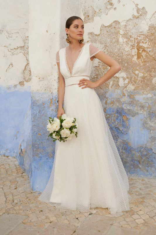 In questa foto una modella, con in mano un bouquet, indossa un abito da sposa 2023 dallo stile boho del brand Marylise 