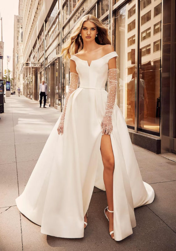 In questa foto la modella posa con un abito da sposa con spacco e guanti in leggero plumetis della collezione Morilee 2023