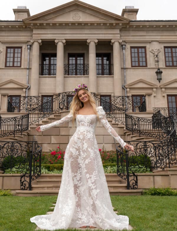 In questa foto una modella indossa un abito da sposa in pizzo floreale, con maniche lunghe, della collezione Reem Acra 2023